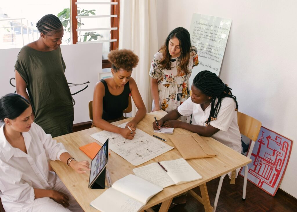 Groupe de femmes collaborant autour d’un projet numérique sur table avec plans, ordinateur et tableau blanc